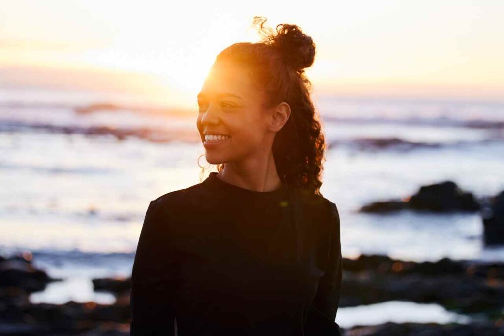 A young African American woman stands in front of the sunset on a beach.