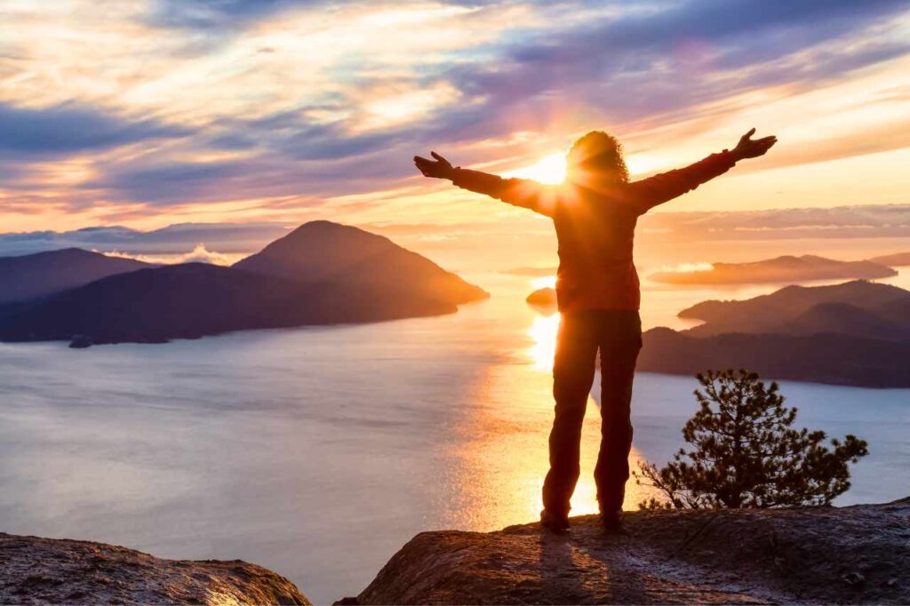 A woman stands on top of a woman overlooking a large body of water at sunset. Her arms are outstretched as she enjoys the view. 