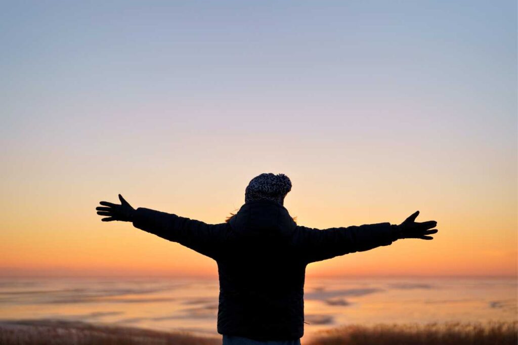 A woman stands in front of a beautiful sunset sky with her arms outstretched.