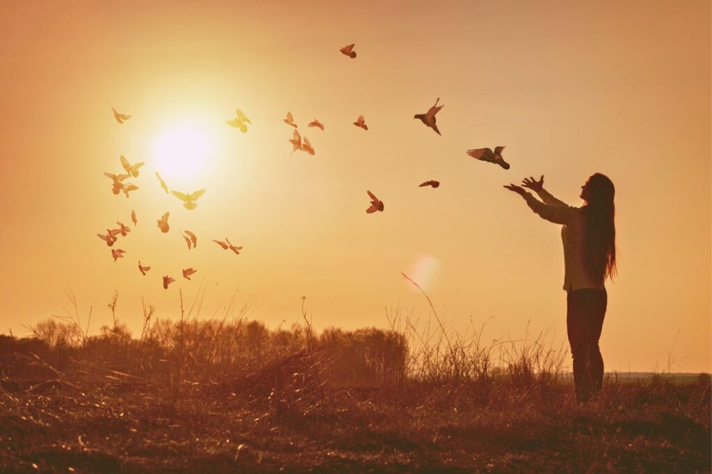 A woman with long hair releases birds, symbolizing hope and freedom, while standing on a hill at sunset.