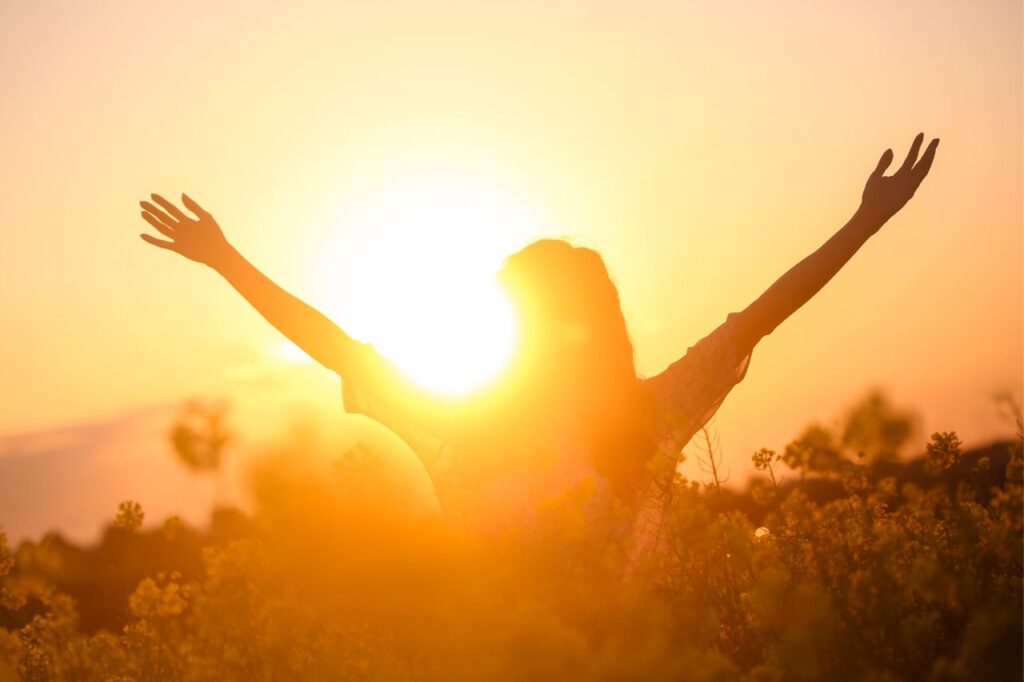 A woman holds out outstretched arms with joy in front of a bright sunset.