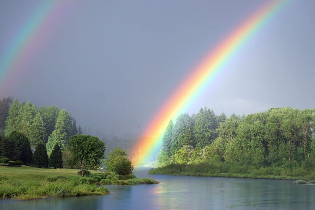 A double rainbow appears over a calm lake with green trees.